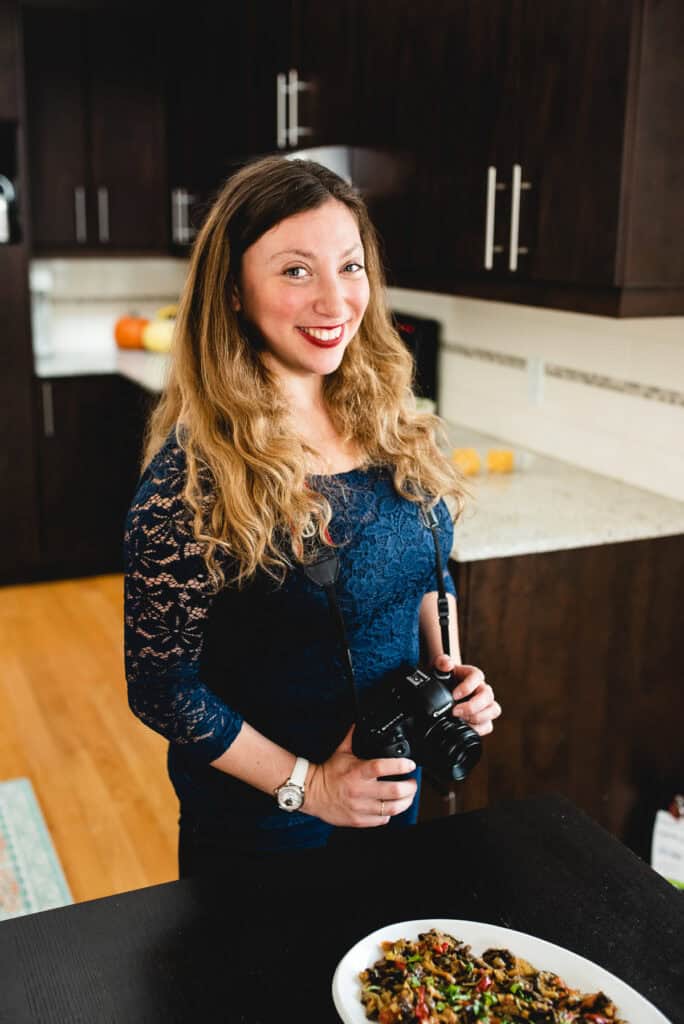 A woman with long hair stands in a kitchen, her camera poised to capture a moment. She's wearing a navy lace top and smiling over a colorful bowl of food. This vibrant scene is set against dark cabinets and light countertops, blending perfectly into what feels like our "About Us" story.