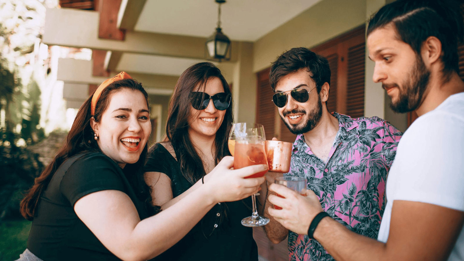 Four people are standing in a casual outdoor setting, raising glasses in a toast. Two women and two men are smiling, wearing casual attire including sunglasses. They appear to be enjoying a drink together near a building with a lantern hanging above.