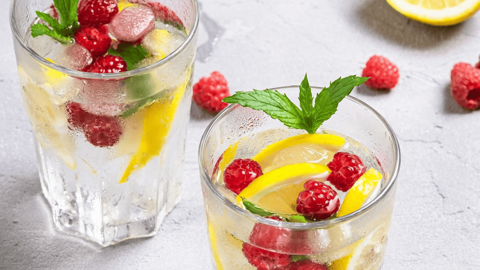Two glasses of refreshing lemonade, reminiscent of infused water recipes, feature lemon slices, raspberries, and mint leaves on a gray surface. Ice cubes chill the mix as extra raspberries and a lemon wedge are artfully scattered around.