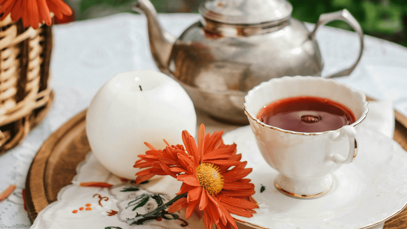 A tea set on a table features a silver teapot, a teacup filled with tea, an orange flower, and a white candle placed on a wooden tray with a patterned cloth. Inspired by Teas Around the World, this elegant display evokes the charm of global tea traditions. A woven basket is partially visible on the left.