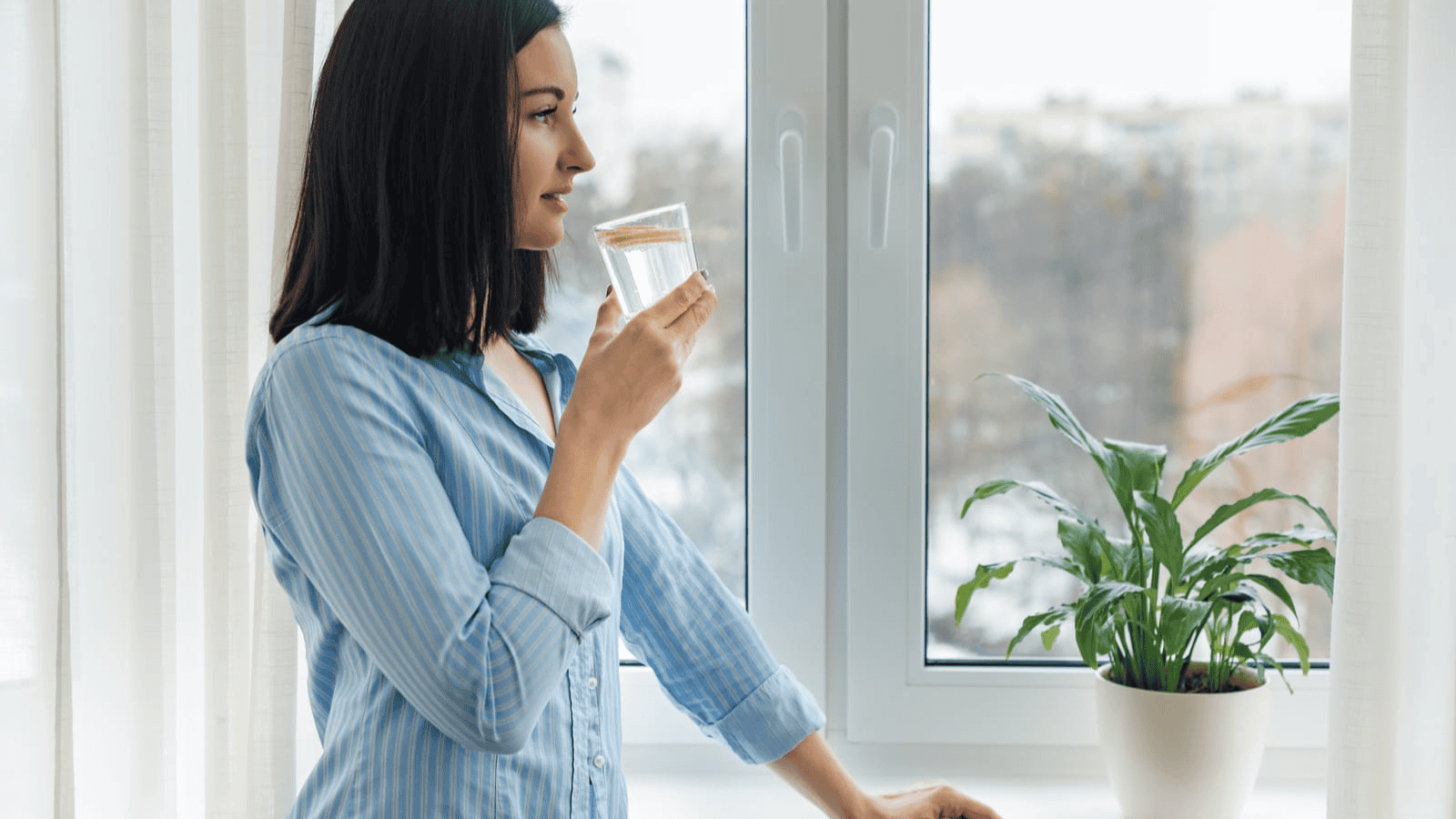 A woman with shoulder-length dark hair stands by a window, looking outside. She is holding a glass of water with a slice of lemon. She is wearing a light blue button-up shirt and is positioned near a potted plant.