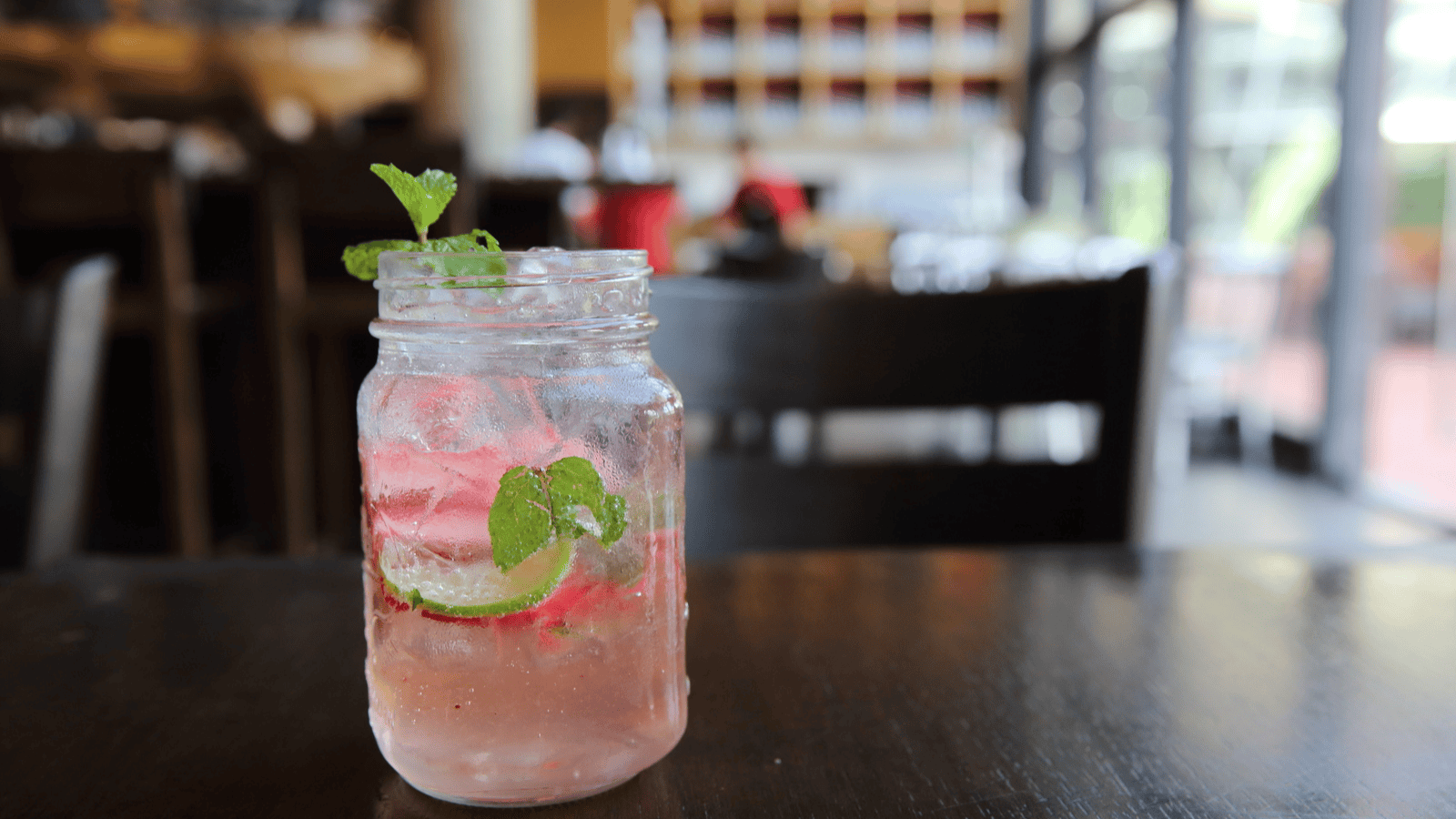 A mason jar filled with a pink-infused water recipe, garnished with lime slices and mint leaves, sits on a dark wooden table. The background captures a blurred view of a cozy cafe with chairs and tables.