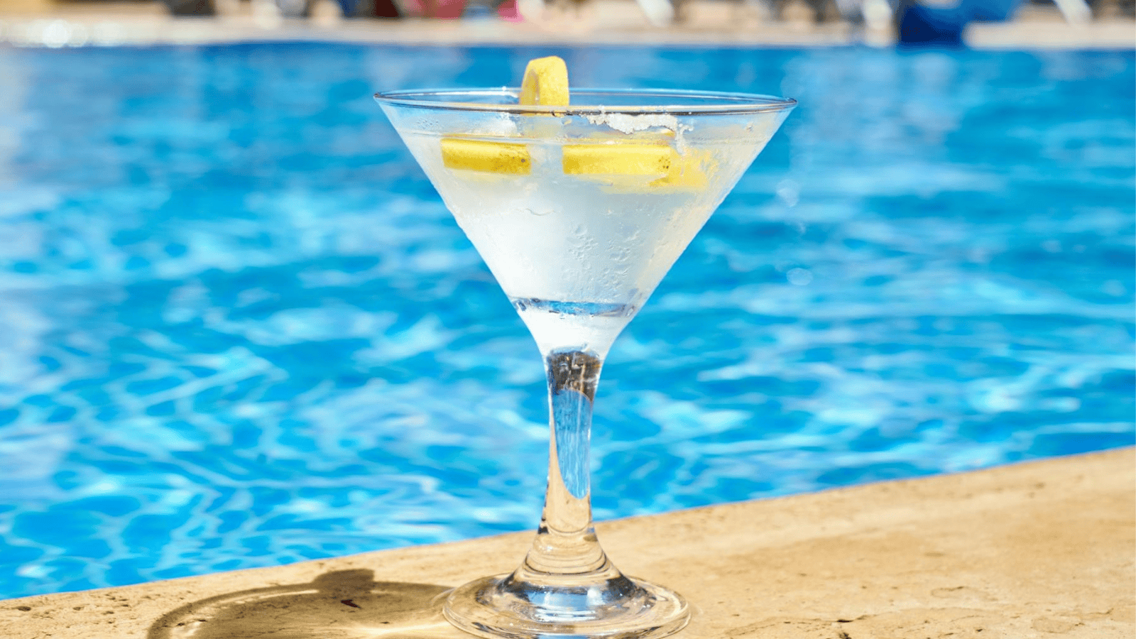 A martini glass filled with a chilled clear liquid and garnished with lemon wedges sits on the edge of a swimming pool. The pool water is a bright blue, and the background is out of focus.