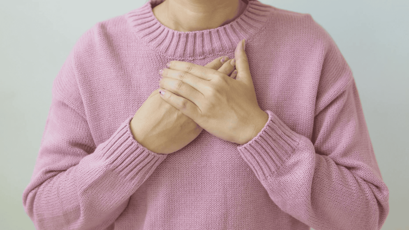 A person wearing a pink sweater holds their hands over their chest, perhaps pondering the subtle signs of alcohol intolerance.