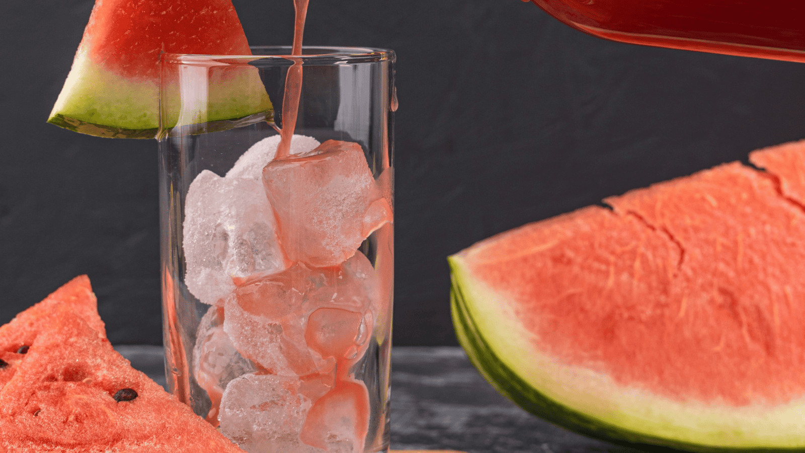A glass brimming with ice cubes is being poured with refreshing watermelon juice, reminiscent of infused water recipes. A juicy watermelon slice adorns the rim, while more vibrant slices rest on the dark surface in the background.