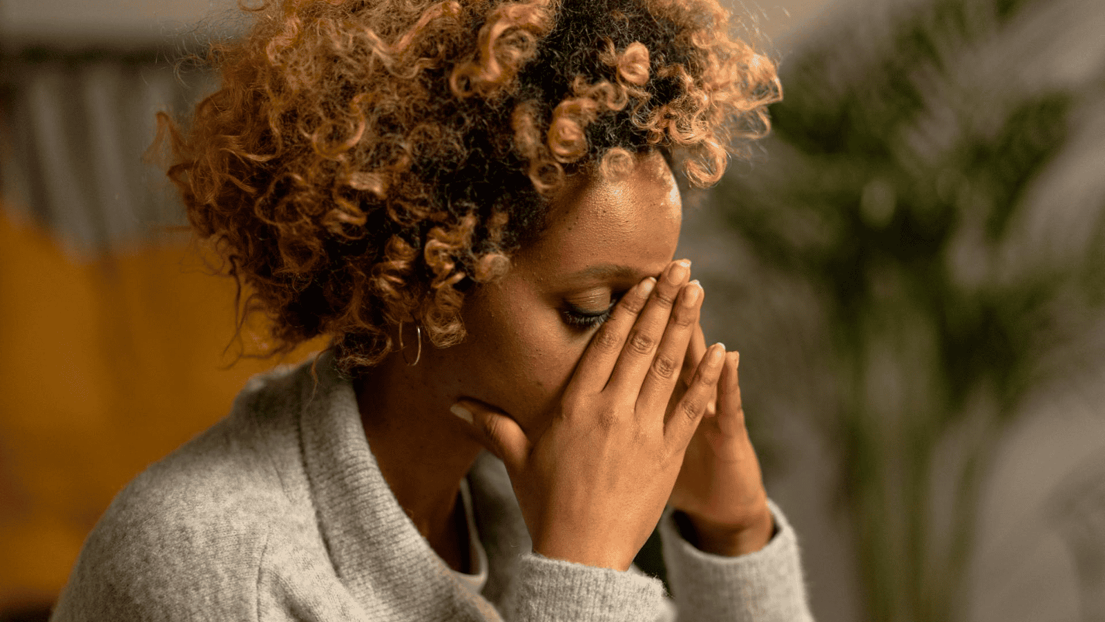 A person with curly hair holds their face in their hands, appearing stressed or frustrated, perhaps contemplating alcohol intolerance signs. They are wearing a gray sweater, and the background is softly blurred with warm lighting and a glimpse of a plant.