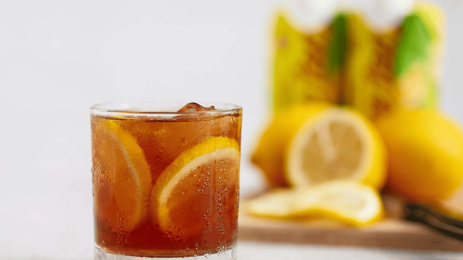 A glass of iced tea with lemon slices sits invitingly in front of blurred lemons and iced tea cartons. Garnished with ice cubes, condensation on the glass hints at a refreshing cold drink, perfect for exploring Teas Around the World.