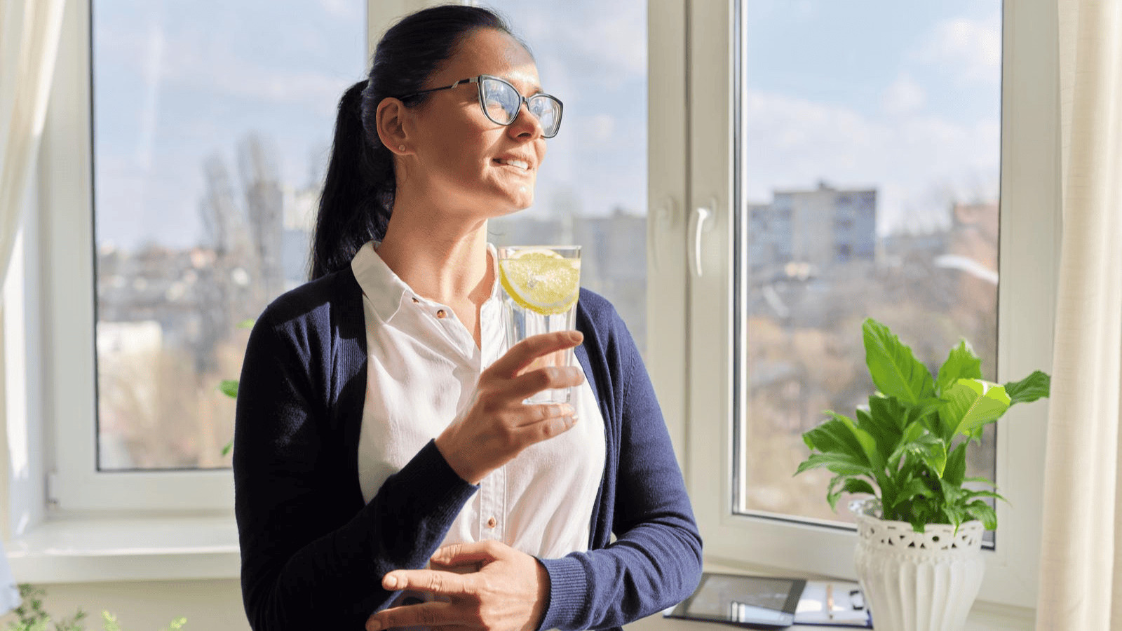 A woman wearing glasses and a navy cardigan holds a glass with a lemon slice. She stands near a window with a view of buildings and a clear sky. A green potted plant is on a table beside her.