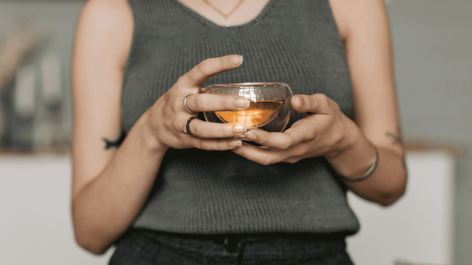 A person in a sleeveless gray top holds a round glass cup with a lit candle inside, reminiscent of evenings spent enjoying Teas Around the World. Multiple rings adorn their fingers, and a tattoo is visible on their right arm. The background is blurred.