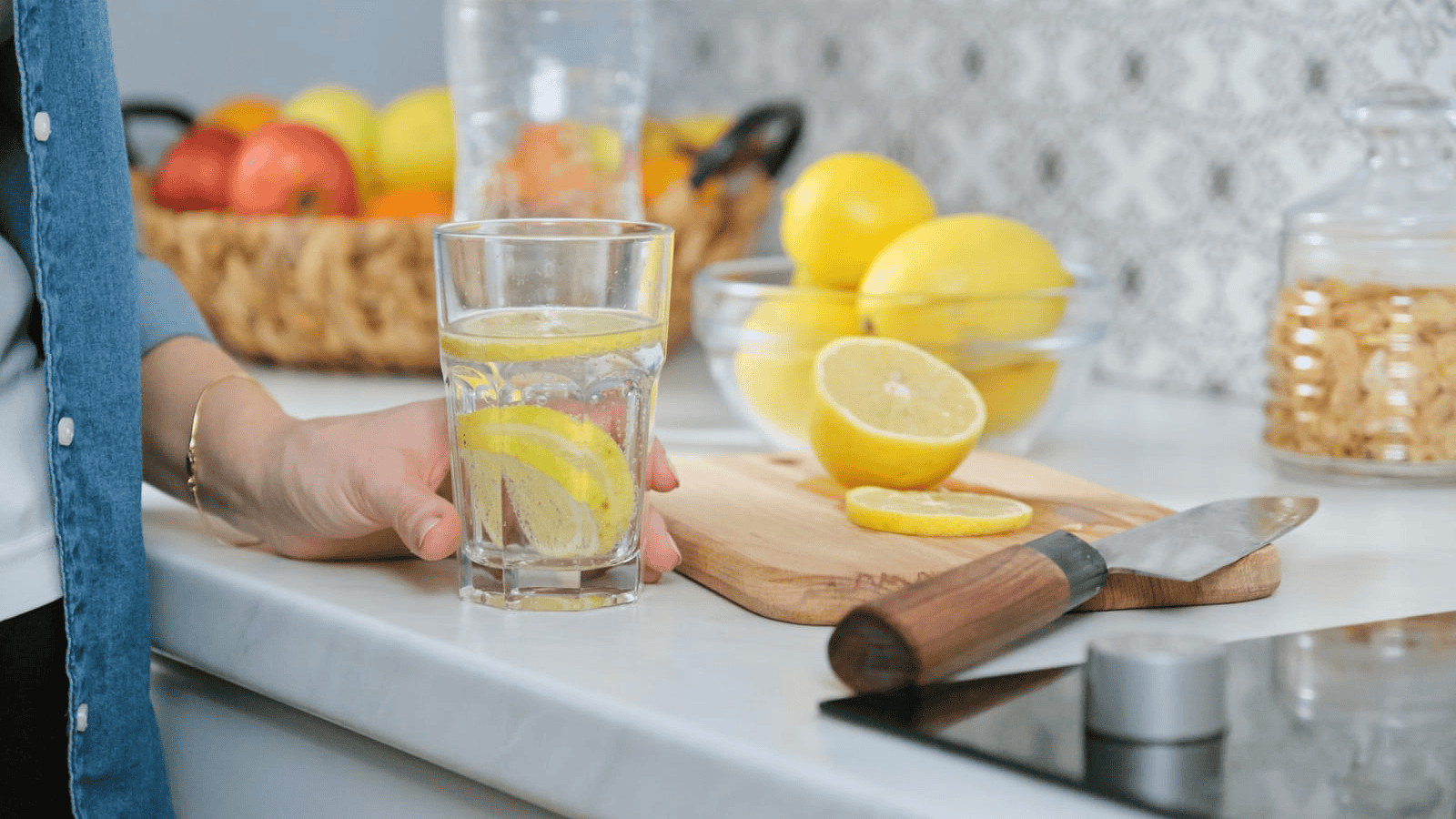 A person holds a glass of water with lemon slices on a countertop. A cutting board with halved and sliced lemons, a knife, and a jar of cookies are also on the counter. In the background, a basket with assorted fruits is visible.