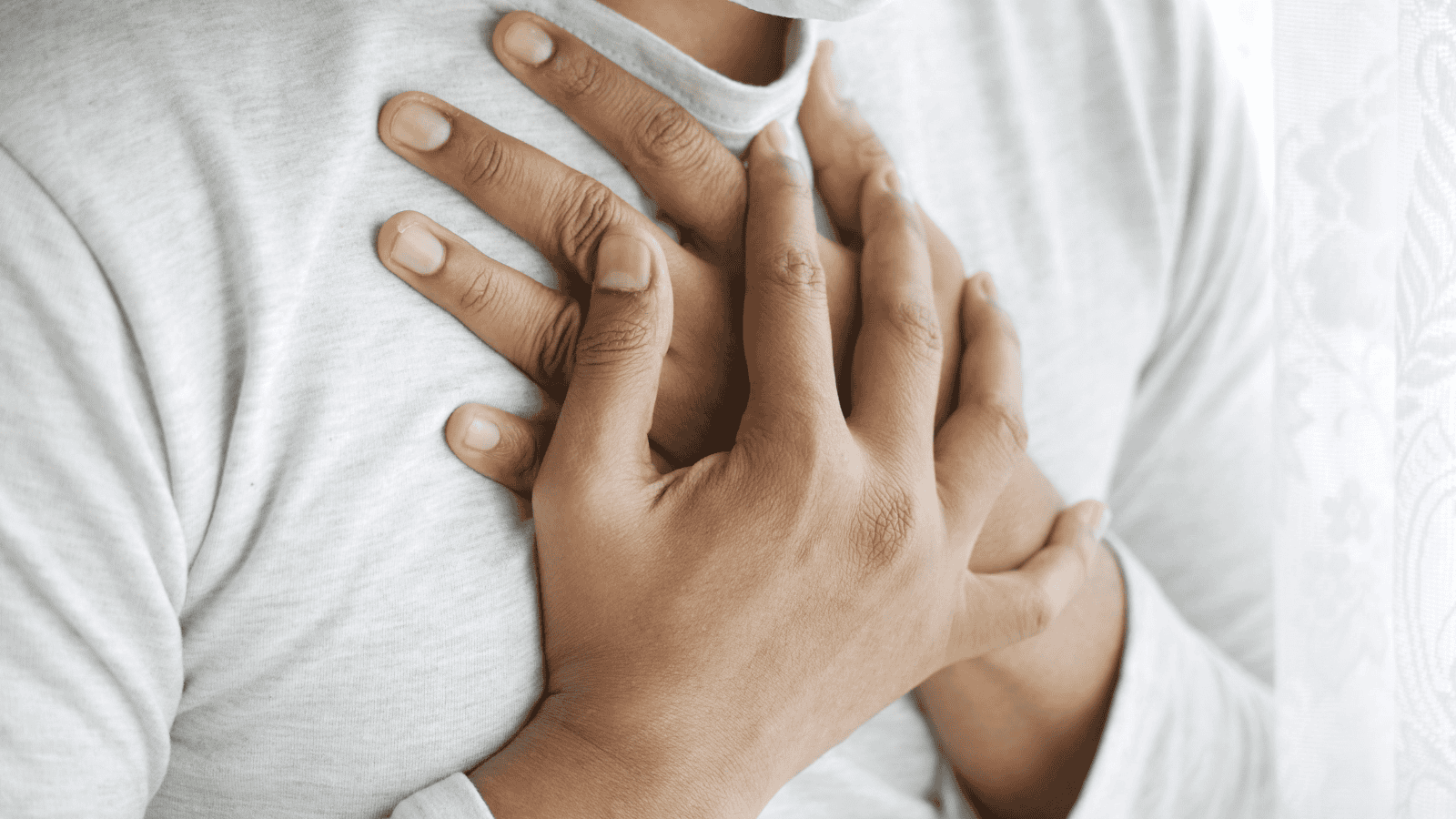 A person wearing a light gray long-sleeve shirt places both hands over their chest, conveying a gesture that could imply pain or emotion—perhaps even hinting at alcohol intolerance signs. The background is softly blurred.