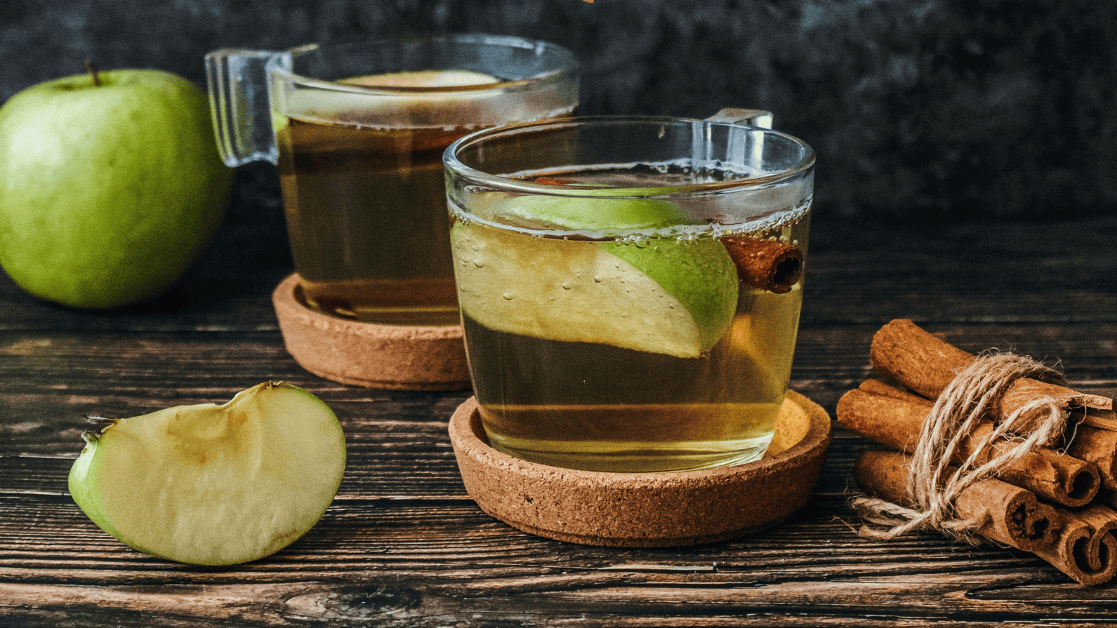 Two clear glass mugs brimming with a spiced apple drink, each adorned with an apple slice and a cinnamon stick, are nestled on wooden coasters. Nearby, a fresh green apple and a bundle of cinnamon sticks rest on the rustic wooden surface, evoking thoughts of creative infused water recipes.