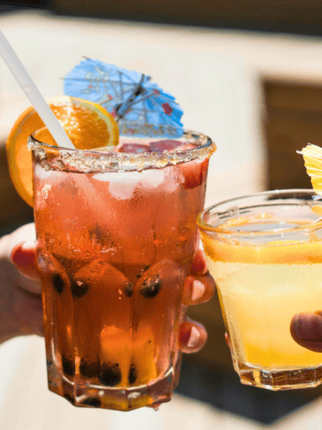 Two people clinking glasses of cocktails, one with a pink beverage garnished with an orange slice and a tiny umbrella, and the other with a yellow drink also topped with a small umbrella. The background is a sunny outdoor setting with wooden deck seating.