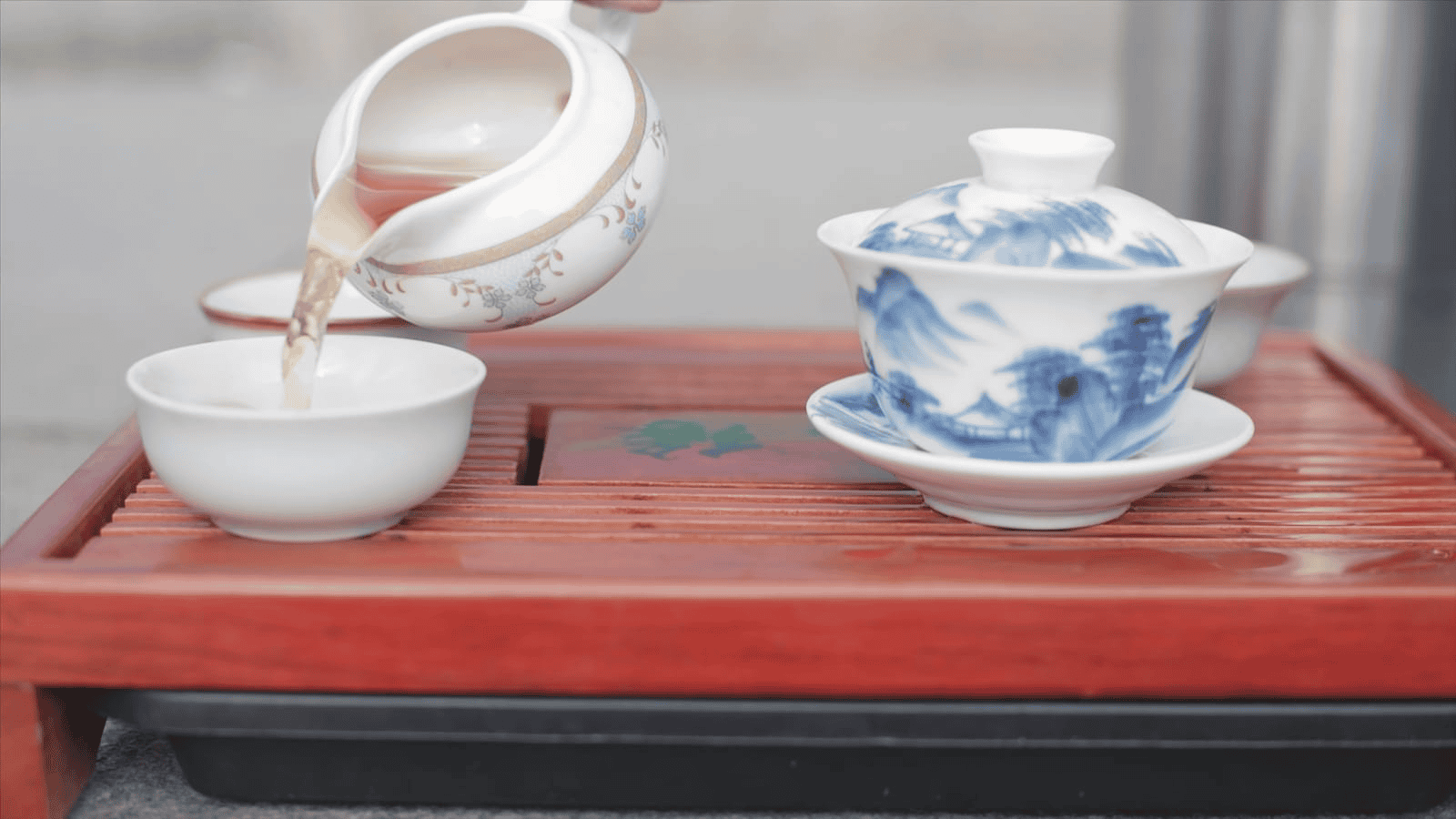 A hand pours fragrant tea from a white and blue teapot into a matching teacup on a wooden tray, evoking the rich traditions of Teas Around the World. Another lidded teacup sits gracefully beside it.