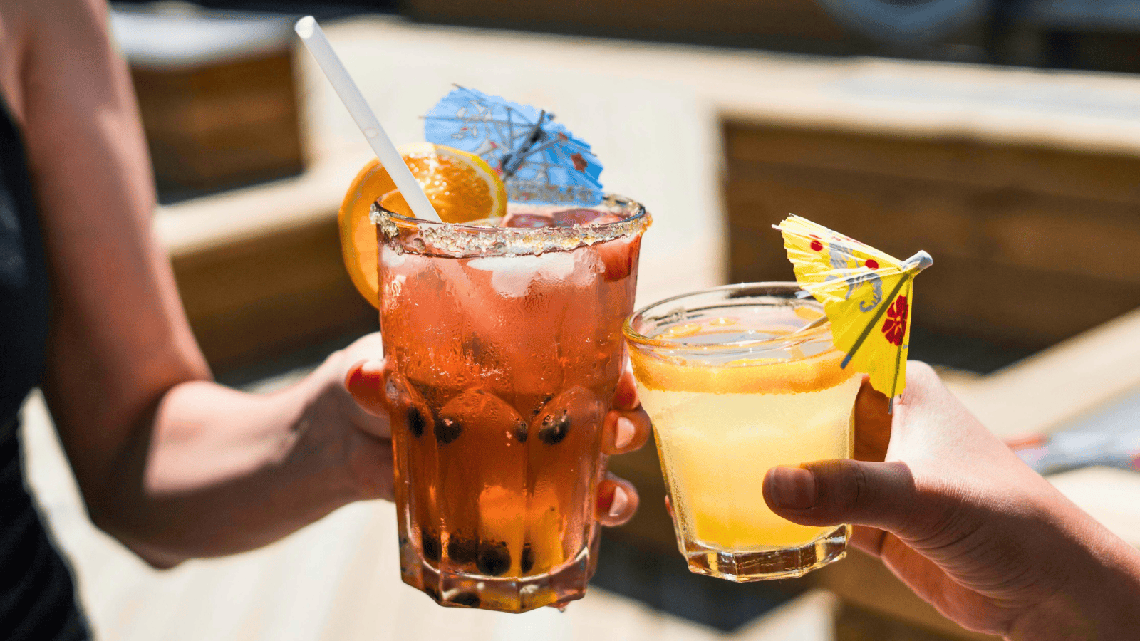 Two people clinking glasses of cocktails, one with a pink beverage garnished with an orange slice and a tiny umbrella, and the other with a yellow drink also topped with a small umbrella. The background is a sunny outdoor setting with wooden deck seating.