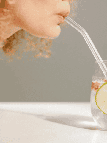 A person with curly hair sips from a clear glass of refreshing infused water, featuring ice, a slice of lime, and a strawberry. The glass rests on a white surface as the person uses a transparent straw. The background is a neutral shade.