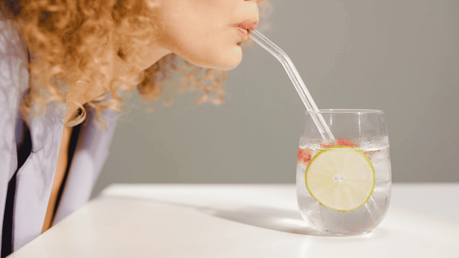 A person with curly hair sips from a clear glass of refreshing infused water, featuring ice, a slice of lime, and a strawberry. The glass rests on a white surface as the person uses a transparent straw. The background is a neutral shade.