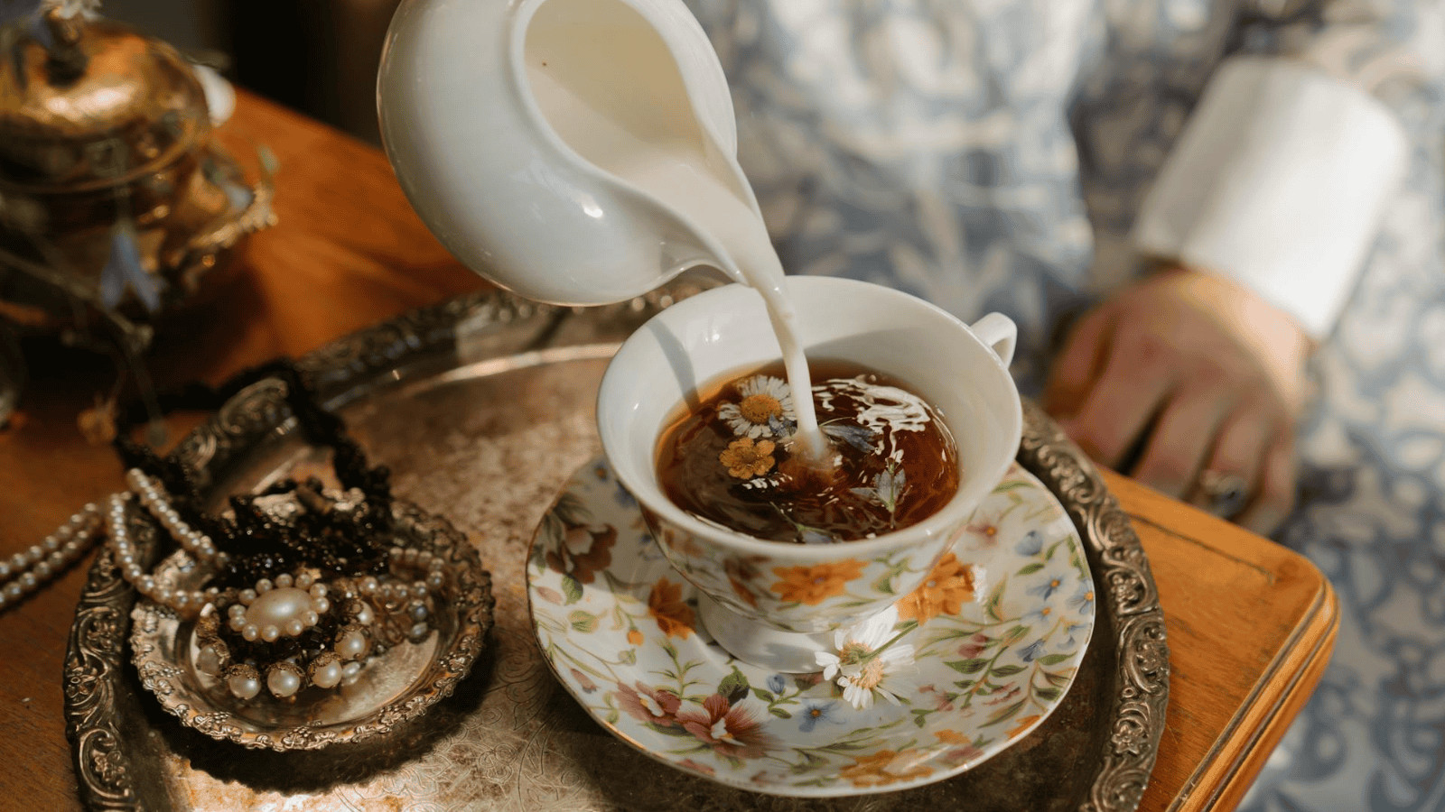 A person is pouring milk into a cup of tea that is placed on a decorative tray. 