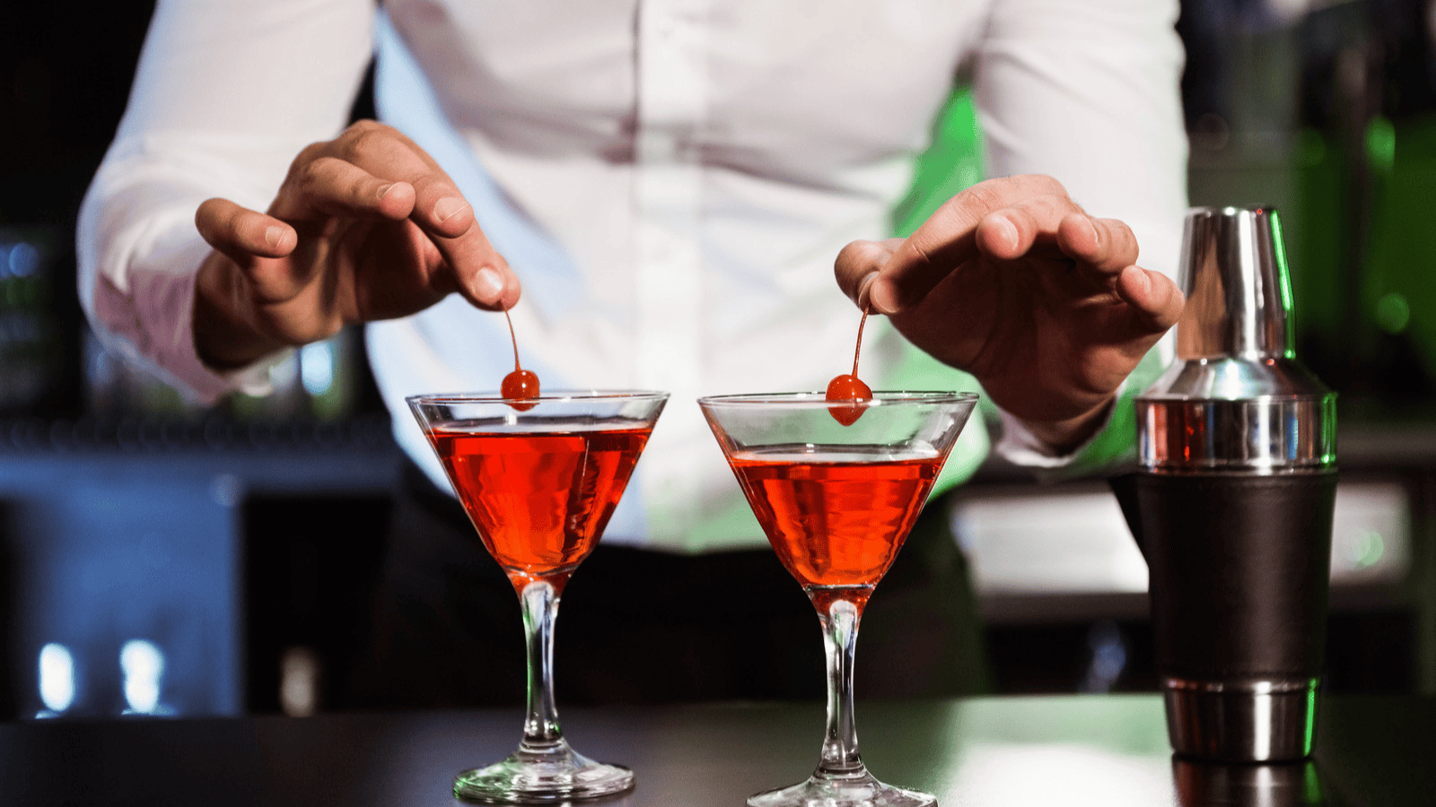 A bartender in a white shirt is placing cherries on top of two red cocktails in martini glasses. A silver shaker with a black grip stands on the bar counter to the right. The background is out of focus with dim lighting.