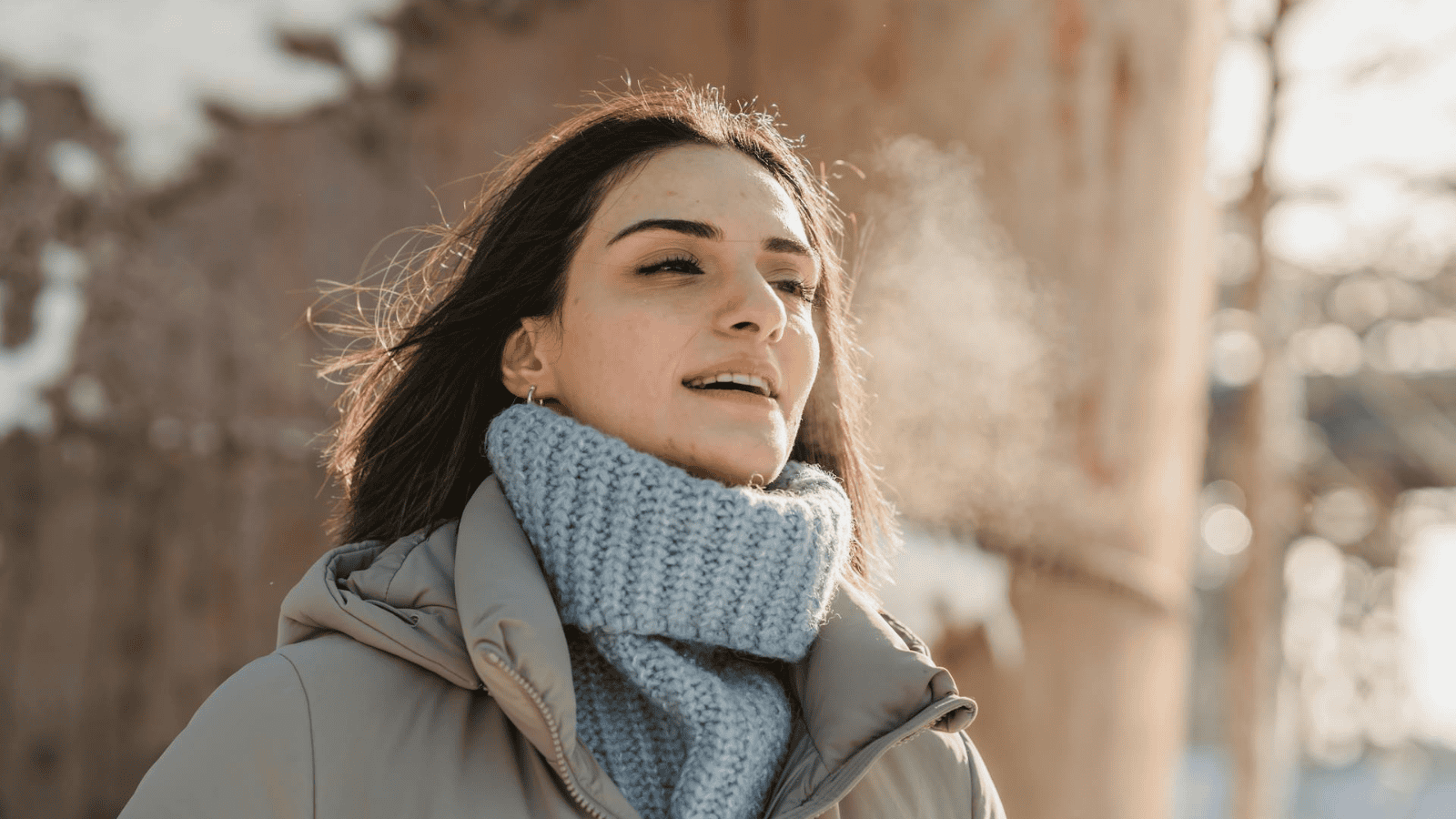 A woman with dark hair and a gray scarf is standing outdoors on a cold day. She is wearing a light-colored coat and visible condensation of her breath suggests it is cold. In the background, there is a blurred, possibly metallic structure.