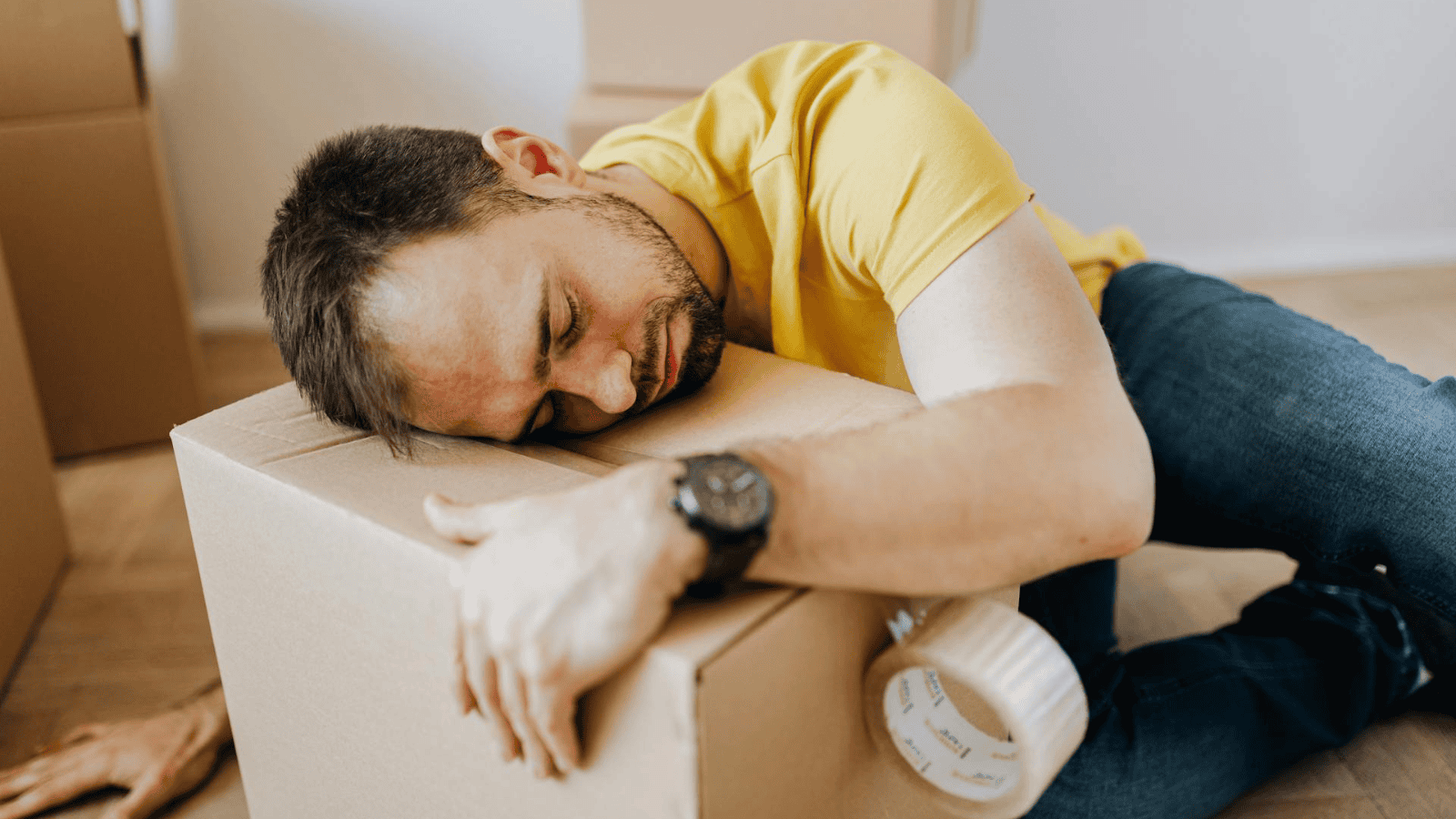 A man in a yellow shirt and jeans lies on the floor, his head resting on a cardboard box. He appears asleep or exhausted, perhaps showing alcohol intolerance signs. A roll of packing tape sits beside him.