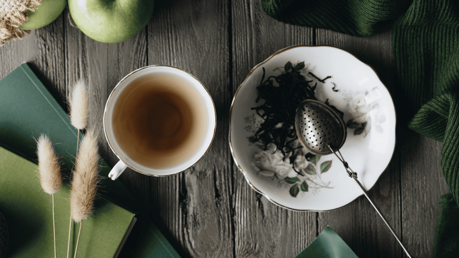 A cup of tea, part of the "Teas Around the World" collection, sits on a wooden table next to a saucer with a tea strainer and loose leaves. Nearby are a green apple, books, dried plants, and fabric, creating a cozy and natural setting.