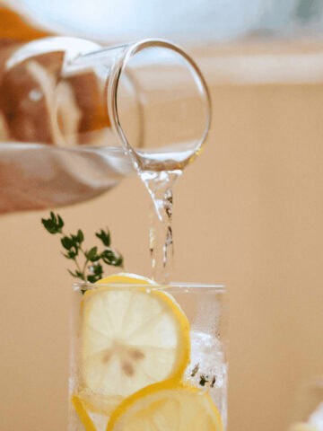 A hand is pouring water from a glass pitcher into a glass filled with ice cubes, lemon slices, and a sprig of thyme. The background is blurred, with soft lighting creating a warm atmosphere.