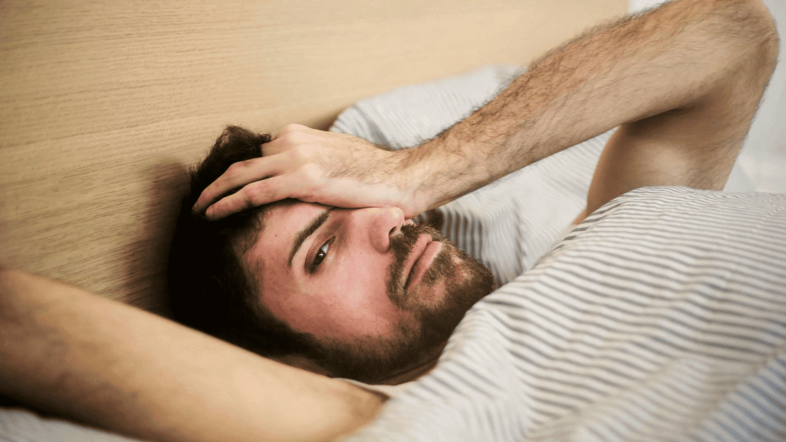 A person lies in bed under a striped blanket, looking exhausted with their hand resting on their forehead. They have facial hair and dark hair, eyes half-open against a wooden headboard—perhaps showing subtle alcohol intolerance signs from the previous night.