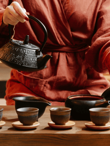 A person in a red robe pours tea from a teapot into four small cups on a wooden table, embodying the essence of teas around the world. The scene unfolds indoors against a warm-toned background, with the dark ceramic teapot and cups adding to the global ambiance.