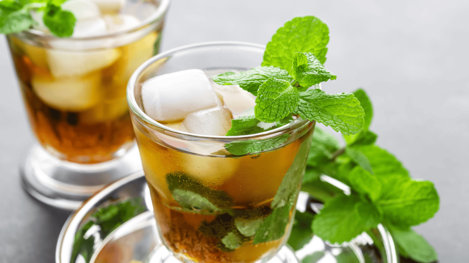 Two glasses filled with a brown iced beverage, garnished with fresh mint leaves and ice cubes, are placed on a reflective surface. Mint leaves are also scattered around the glasses. The background is a blurred grey surface.