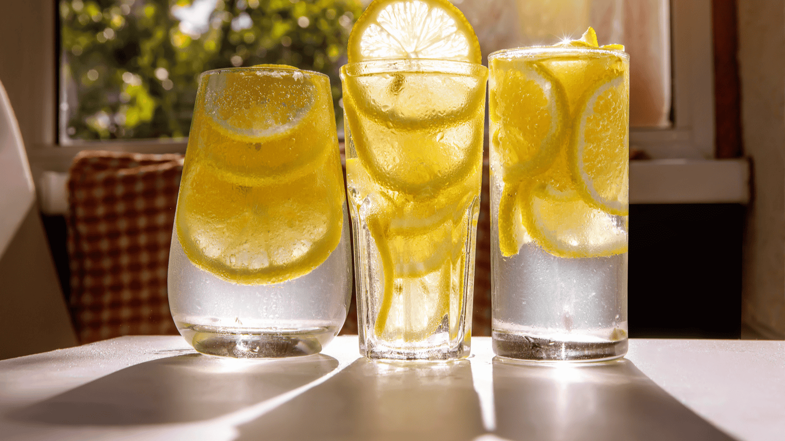 Three clear glasses filled with lemonade, each containing ice cubes and lemon slices, are set on a sunlit surface. The background shows a window with sunlight streaming through, casting shadows and highlights on the glasses.