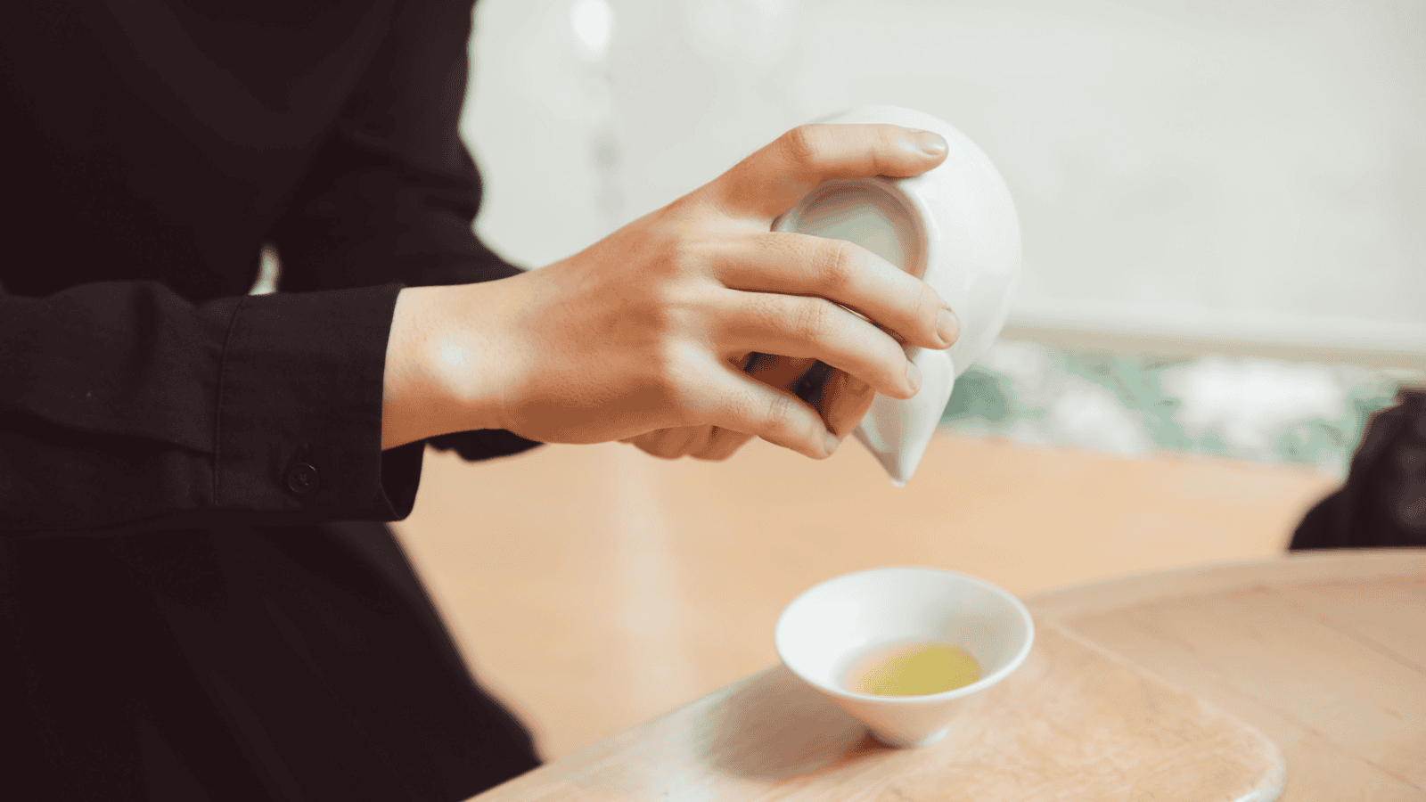 A person dressed in a black outfit is pouring tea from a white teapot into a small white teacup on a wooden surface, evoking the charm of "Teas Around the World." The blurred background suggests an inviting indoor setting.