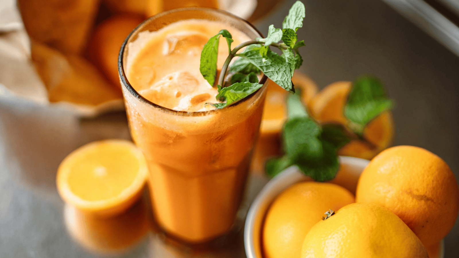 A glass of orange juice with ice and a sprig of mint on top is on a table. Next to it, there are halved and whole oranges along with some mint leaves. In the background, there is a bag filled with more oranges.