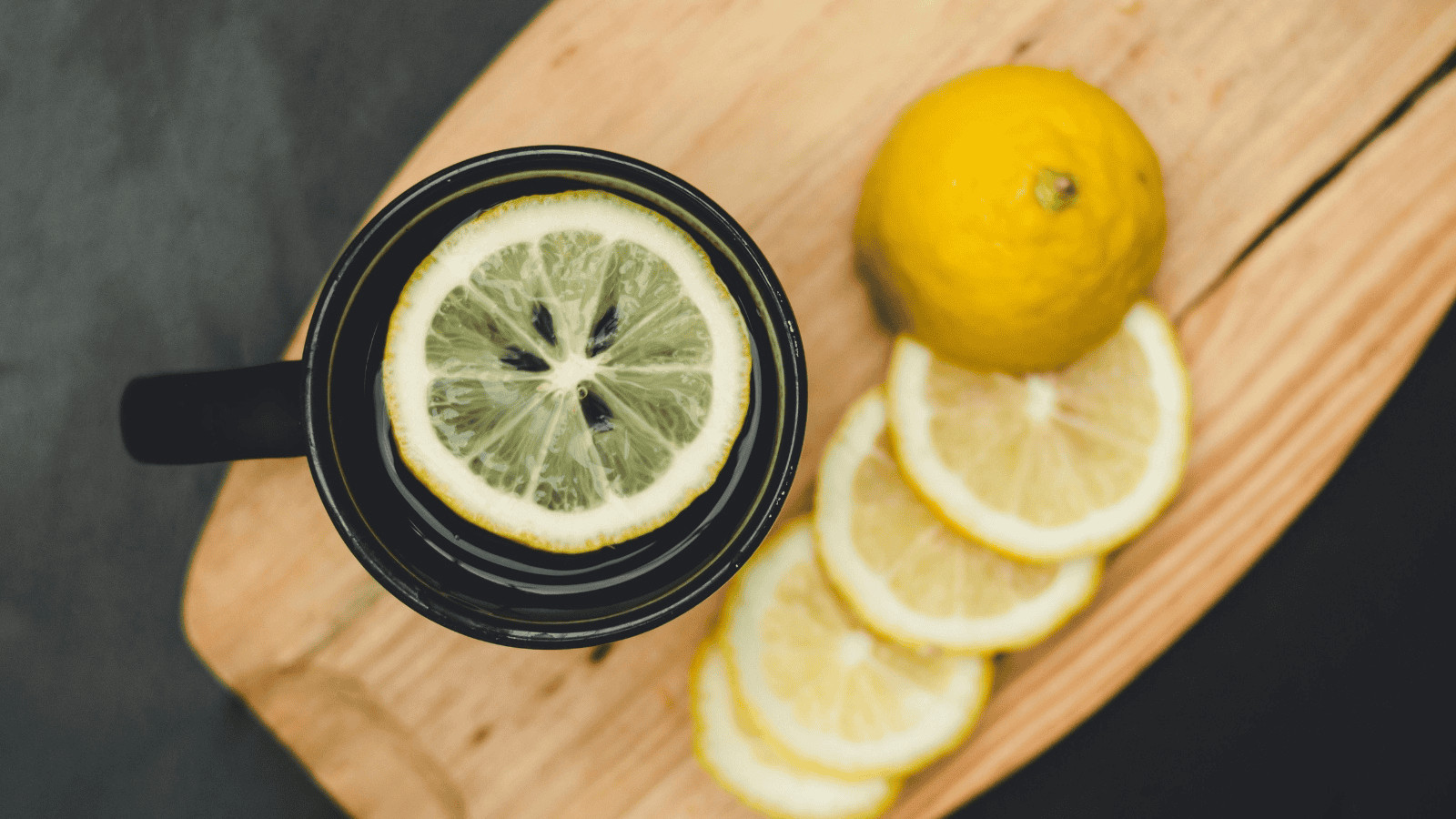 A black cup filled with water and a slice of lemon on top sits on a wooden cutting board. A whole lemon and several slices of lemon are arranged next to the cup on the cutting board.