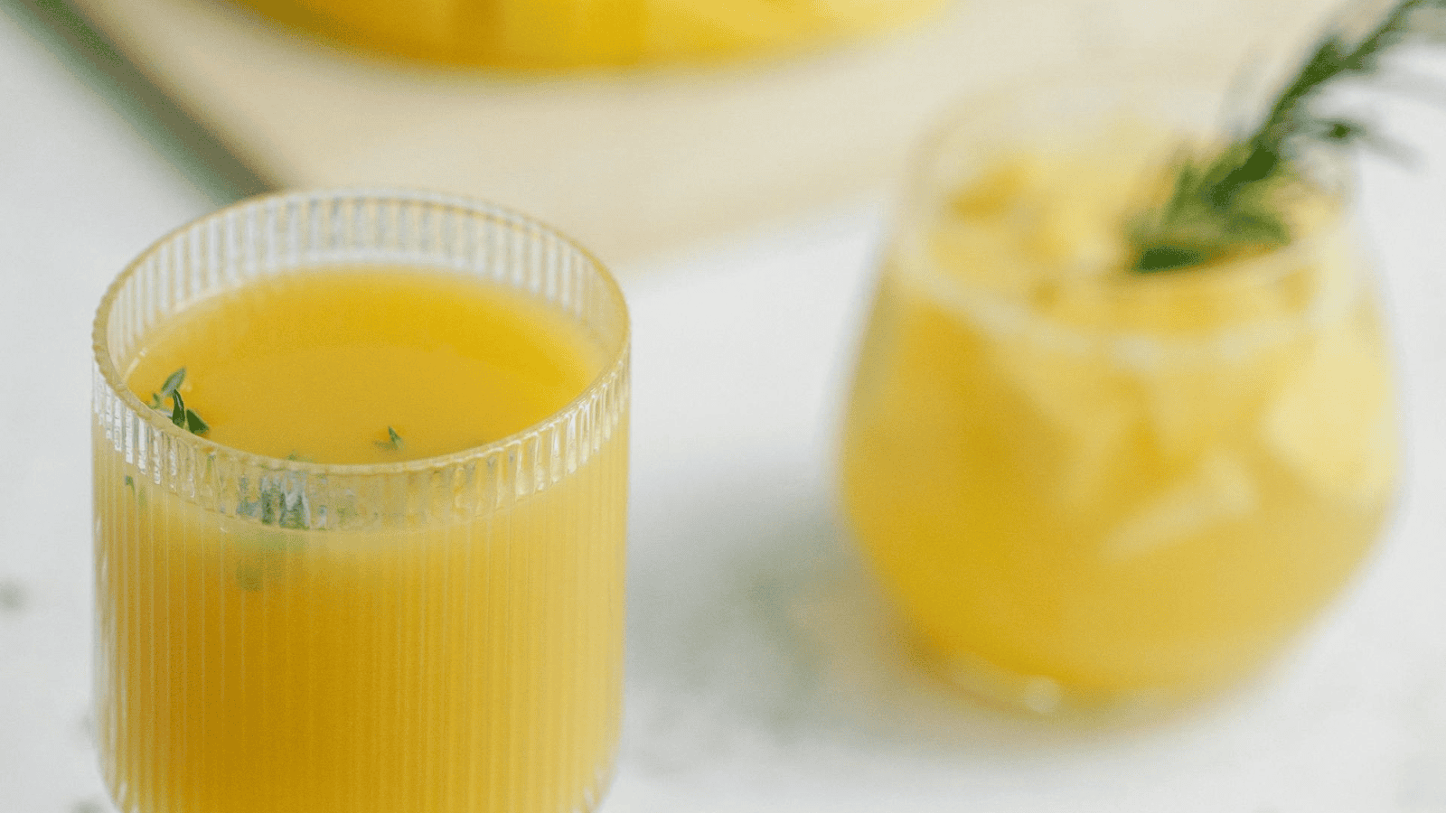 Two glasses containing yellow beverages are on a white surface. The glass on the left has a textured design and is filled with juice, while the glass on the right features infused water recipes with ice cubes, yellow liquid, and a green garnish.