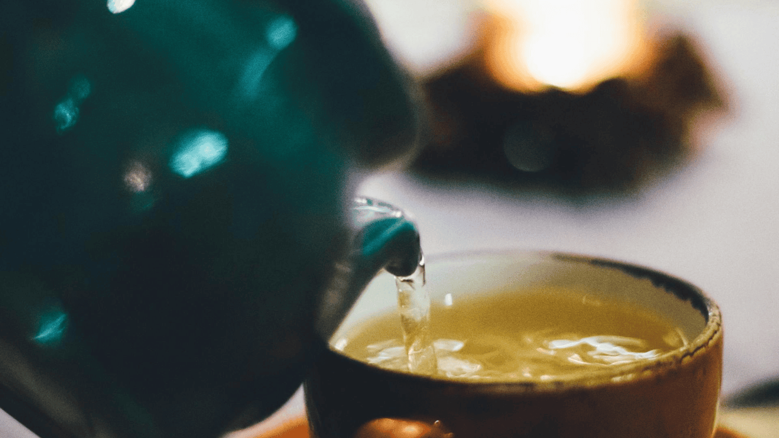 A close-up of a teal teapot from the Teas Around the World collection pours hot liquid into a ceramic cup, creating ripples on the surface. In the blurred background, a softly glowing light suggests a warm, cozy setting.