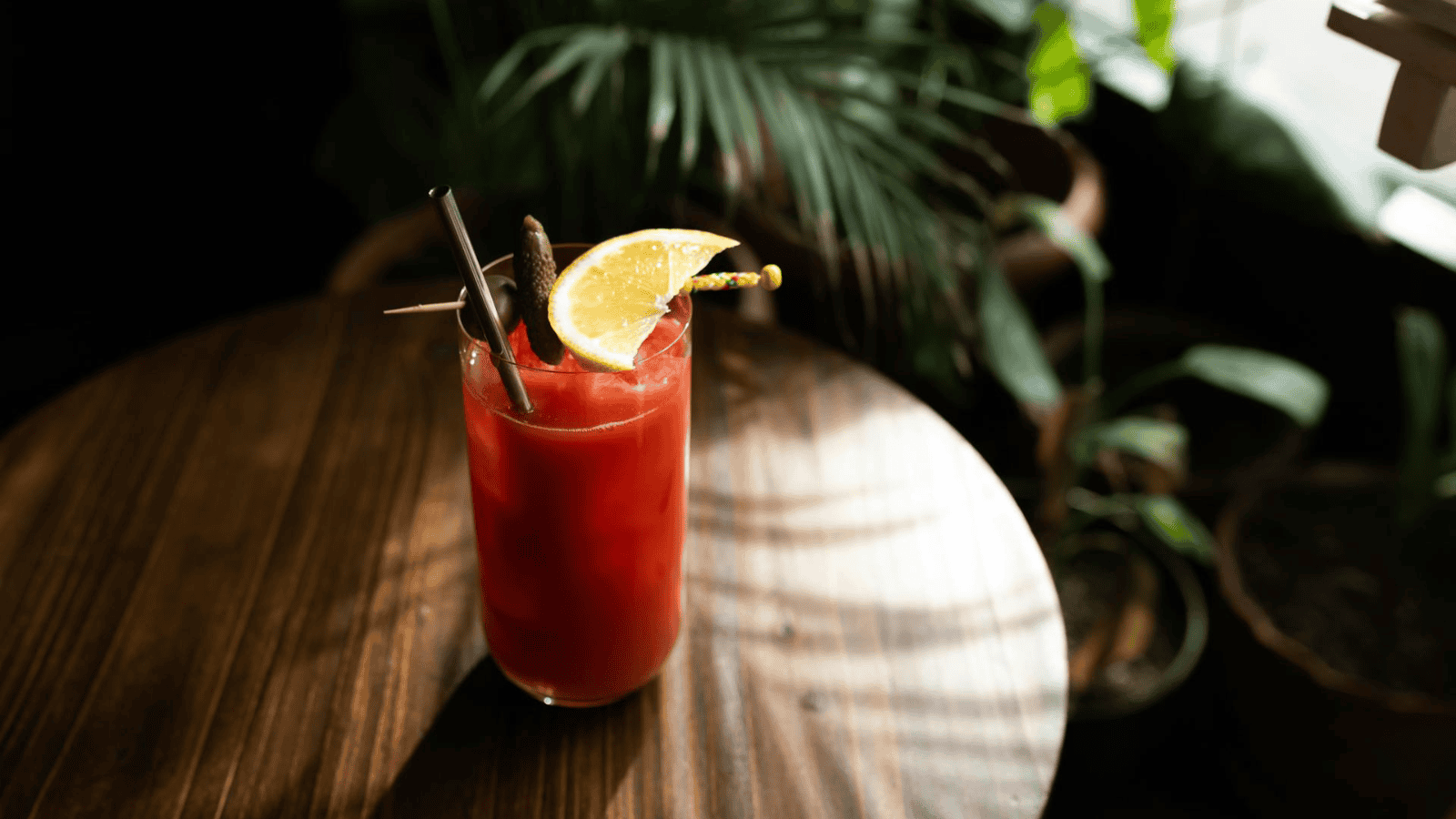 A glass with a red drink, garnished with a slice of orange and a pickled garnish, is placed on a round wooden table. A straw is also inserted in the drink. The background features potted green plants.