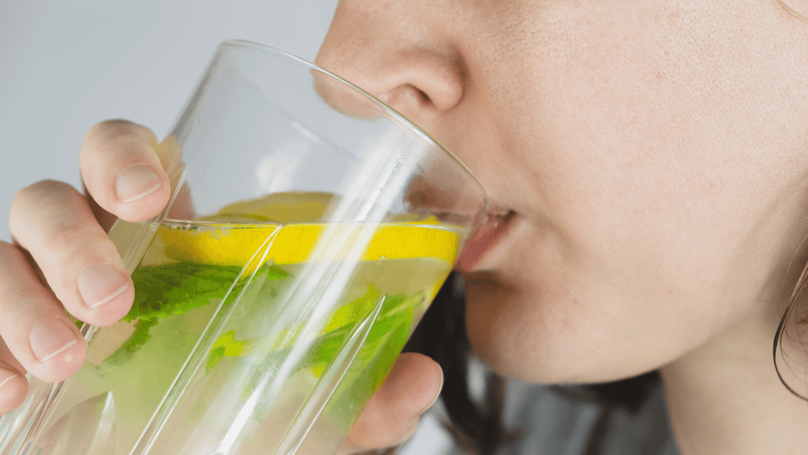 A close-up image shows a person drinking from a glass of water with lemon slices and mint leaves. The person's lips are touching the rim of the glass. Only part of the person's face is visible, focusing on the mouth and chin.