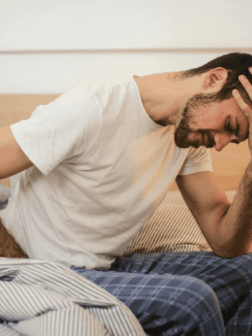 A man in a white t-shirt and blue plaid pants sits on a bed, holding his head with one hand. His tense expression hints at alcohol intolerance signs. The bed has striped bedding, and a small nightstand with a lamp is nearby.