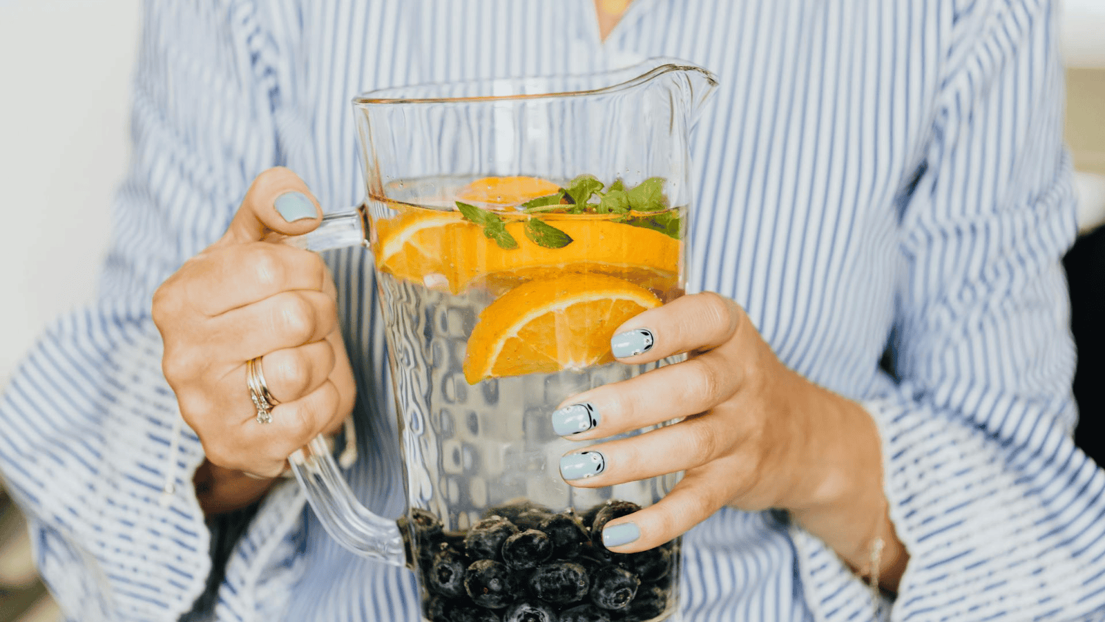 A person holding a clear pitcher of a refreshing infused water recipe with orange slices and blueberries. The individual is wearing a blue and white striped shirt and has light blue nail polish, perfectly complementing the vibrant colors of the fruit-filled drink.