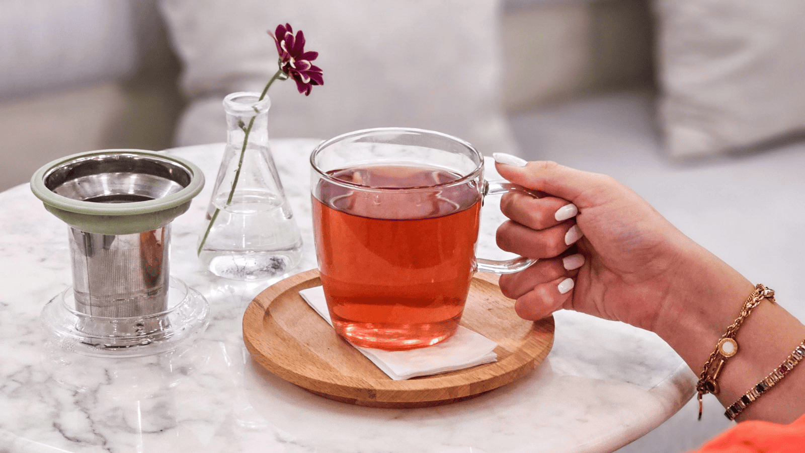 A clear glass mug filled with red herbal tea, reminiscent of blends from "Teas Around the World," sits on a wooden coaster on a marble table. A hand with white nail polish holds the mug. Nearby, a small vase with a single red flower and a metal tea strainer add to the scene's elegance.