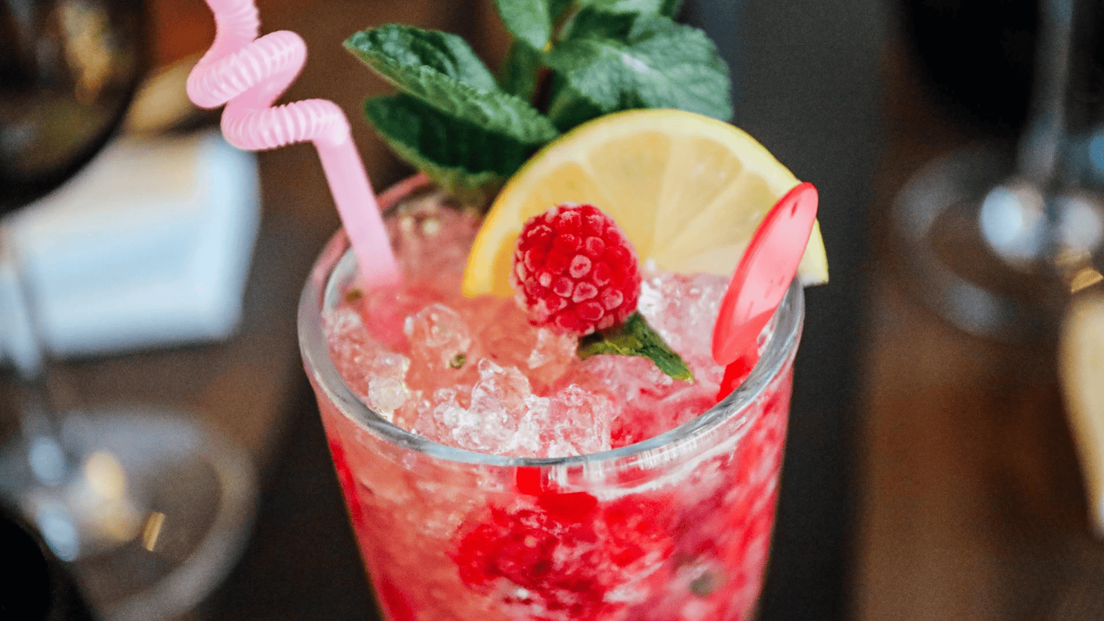 A refreshing pink drink served in a glass filled with crushed ice, garnished with raspberries, a lemon slice, fresh mint leaves, and a pink plastic stirrer. The drink has a light pink straw with a spiral design.