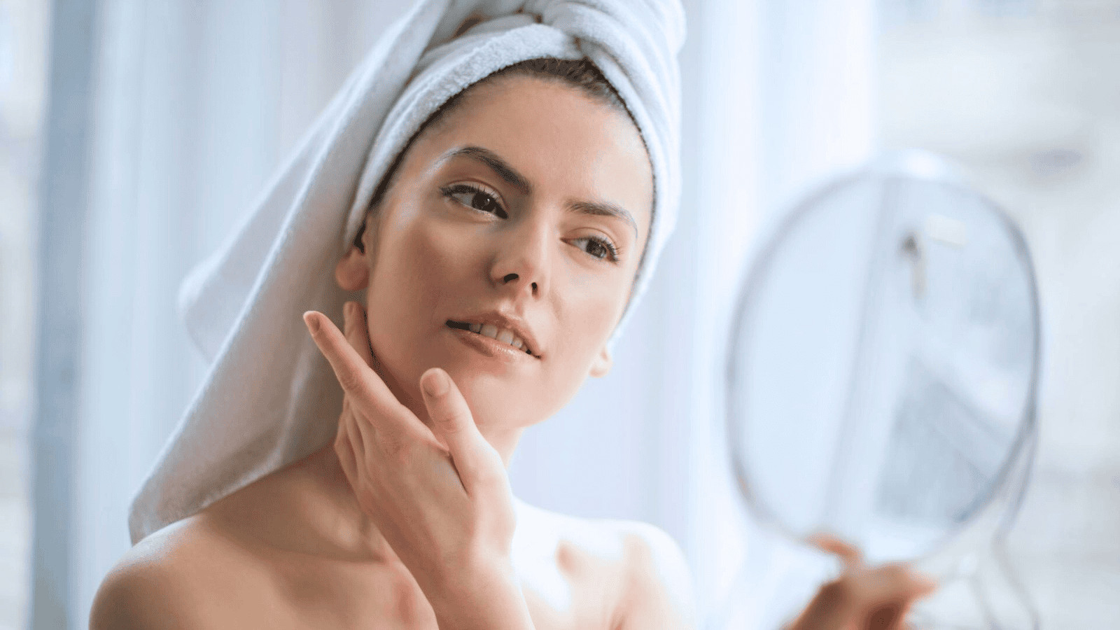 A person with a towel wrapped around their head looks into a handheld mirror while gently touching their cheek. The background is softly blurred, giving a bright and airy feel to the image.