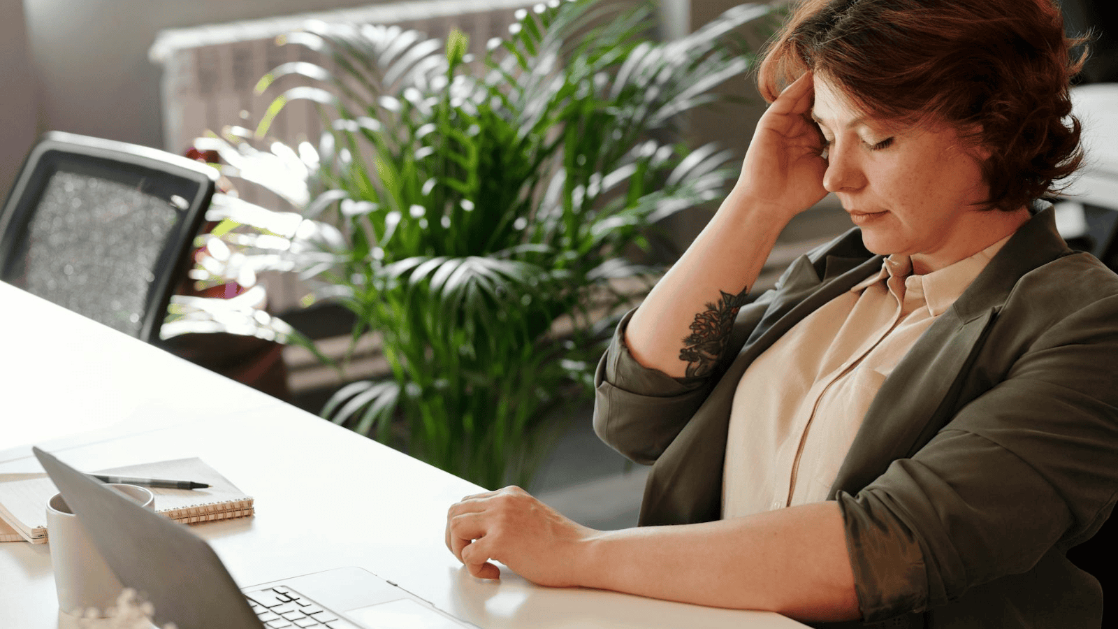 A person with short hair sits at a desk, resting their head on one hand, possibly pondering signs of alcohol intolerance. They appear thoughtful or tired. A laptop is open in front of them, and a notebook with a pen is nearby. A large plant and an office chair are in the background.