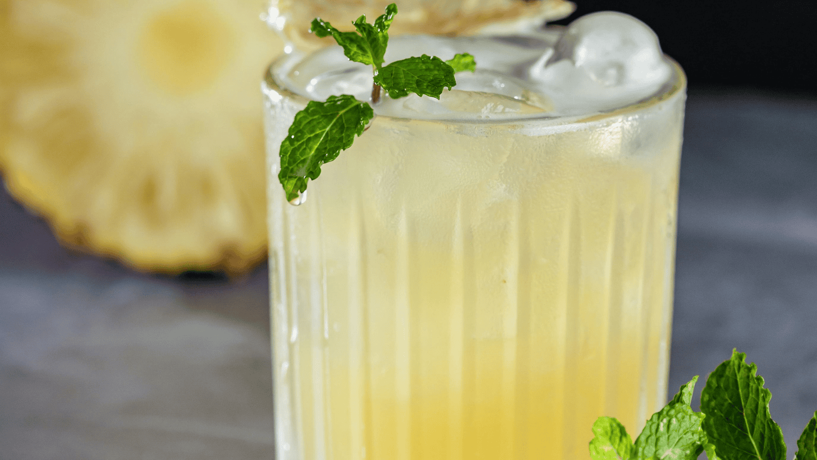 A glass filled with a light yellow drink, topped with ice and garnished with mint leaves. This refreshing blend, reminiscent of infused water recipes, features a slice of pineapple in the background to add a tropical touch.