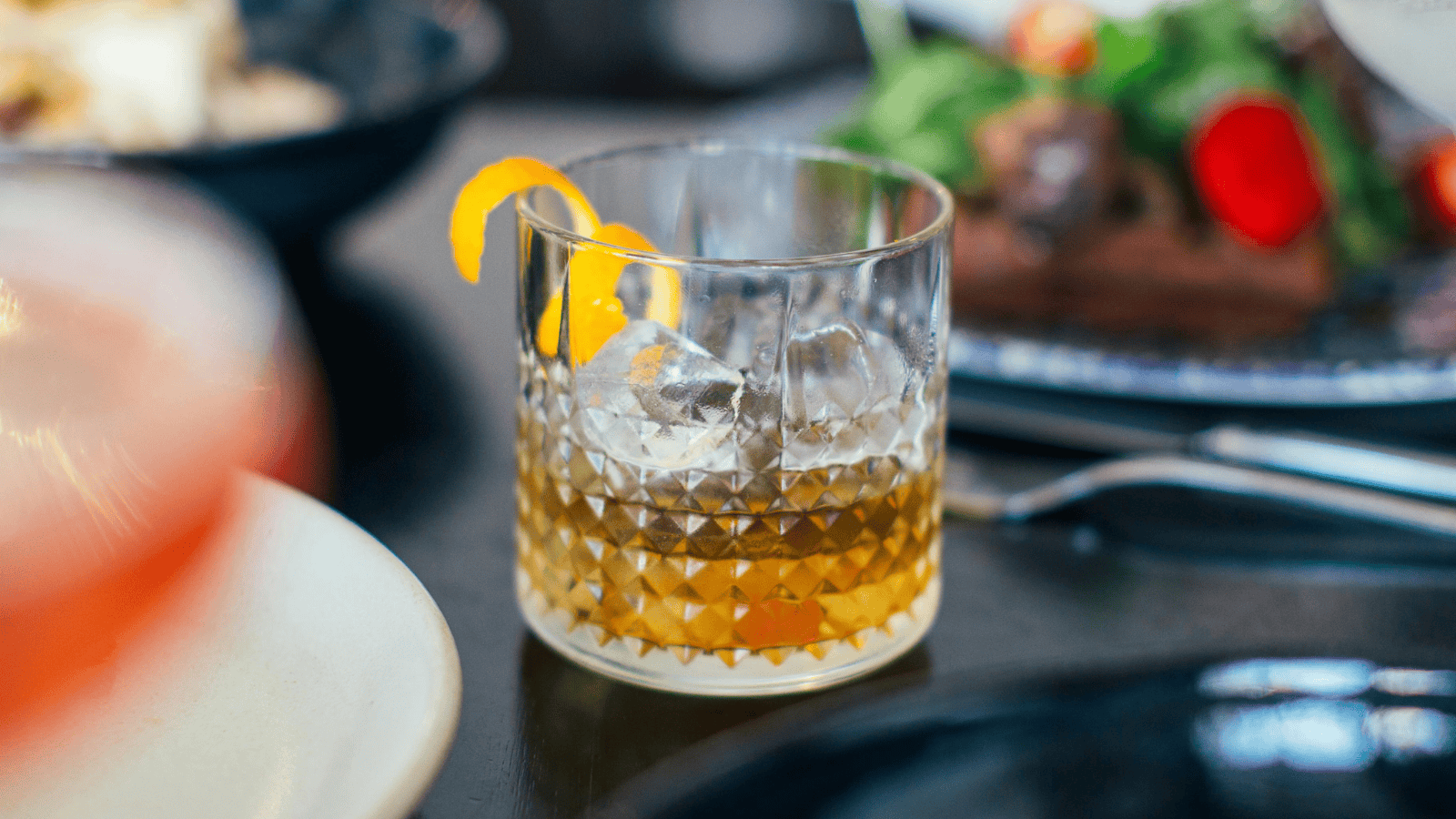 A clear glass with a textured pattern filled with an amber-colored drink, ice cubes, and a twist of orange peel is placed on a table. Plates of food are visible in the background, slightly blurred.