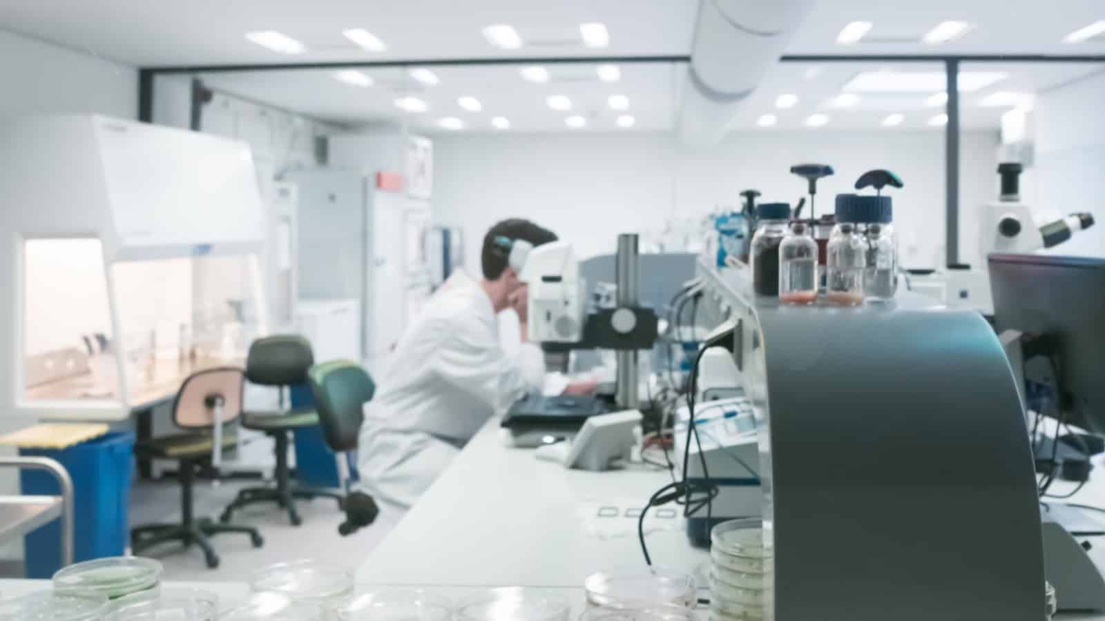 A scientist wearing a white lab coat and headphones is working at a computer station in a laboratory. The workspace includes various scientific equipment, Petri dishes, and bottles. The lab is well-lit with bright overhead lights.