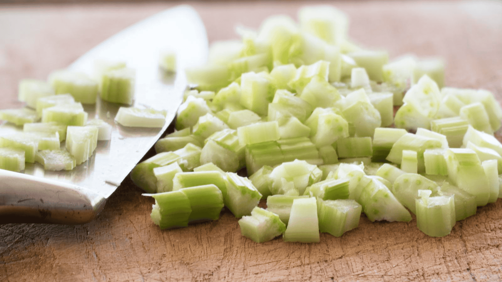 Close-up of diced celery on a wooden cutting board with a stainless steel knife next to it. The celery pieces are irregularly shaped, with the knife partially embedded in the pile. The image focuses on the freshly cut vegetables.
