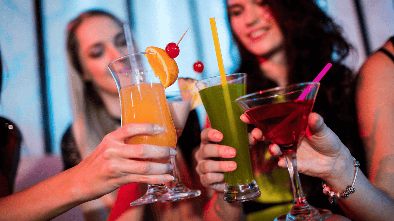 A group of people raise various colorful cocktails in a toast. The drinks include an orange beverage with citrus garnish, a green drink with a cherry garnish, and a red cocktail in a martini glass. The background is blurred.
