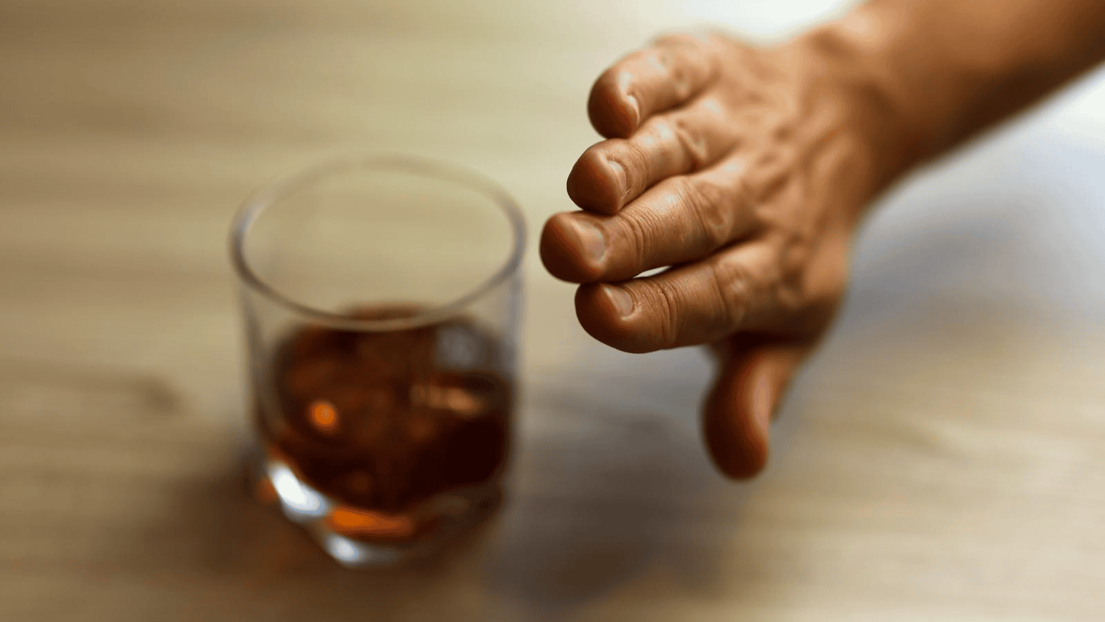 A hand with extended fingers gestures to stop near a glass filled with a dark liquid, possibly an alcoholic drink, placed on a wooden surface.